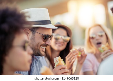 Group Of Friends Sitting On The Beach And Eating Sandwiches During Picnic.