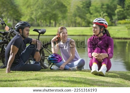 group of friends sitting on the lawn beside the pond after cycling in the park on summer time, three of  cheerful friends enjoy talking together in nature 