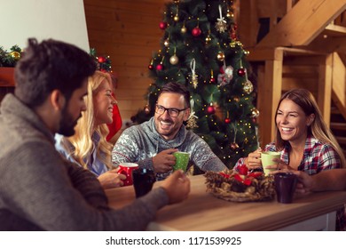 Group Of Friends Sitting On The Living Room Floor Next To A Nicely Decorated Christmas Tree On A Christmas Morning, Drinking Coffee And Having Fun