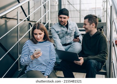 Group of friends sitting on stairs, using social media with smartphones and laptop, smiling and having interesting discussion.