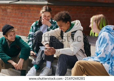 Group of friends sitting on steps of building outdoors and laughing while watching funny video on mobile phone