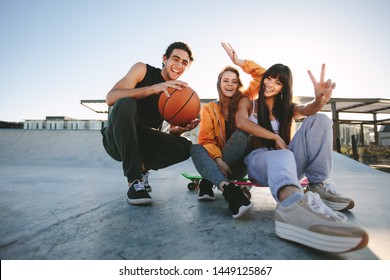Group Of Friends Sitting At Skate Park With Skateboard And Basketball. Male And Female Friends Posing For A Portrait At Skate Park.