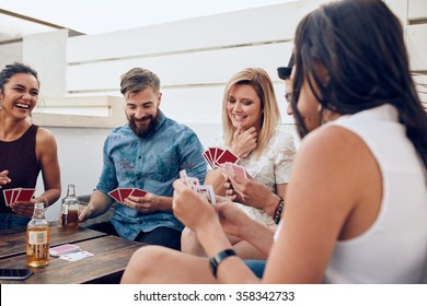 Group Of Friends Sitting At A Wooden Table And Playing Cards. Young People Playing A Game Of Cards During A Party.