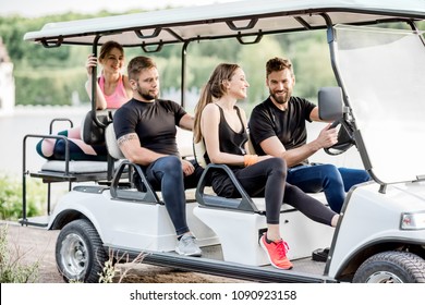 Group Of Friends In Sports Wear Having Fun Driving A Golf Cart Near The Lake