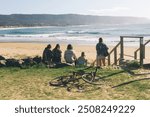 A group of friends stands on the beach, backs to the camera, gazing at the surf. Their silhouettes are framed by the rolling waves and golden light, capturing a moment of shared anticipation and joy.