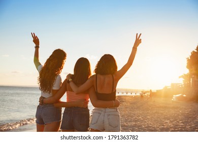 Group Of Friends Staying And Hugging With Raised Arms On The Beach. Travel Or Sea Vacations Concept. Three Young Happy Women In The Summer. View From Back.