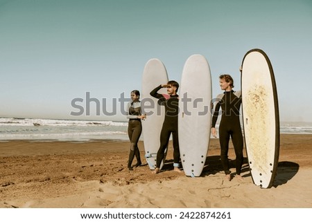 Group of friends surfers in wetsuits standing with surfboards and looks away