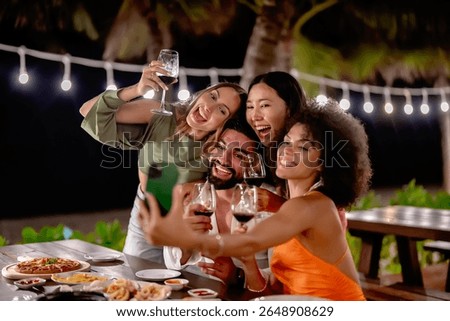 Group of friends taking a selfie while enjoying drinks and dinner at an outdoor tropical restaurant, smiling and celebrating under string lights