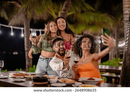 Group of friends taking a selfie while enjoying drinks and dinner at an outdoor tropical restaurant, smiling and celebrating under string lights