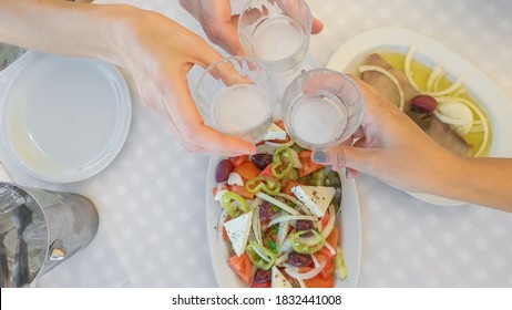 Group of friends toasting together at a restaurant top view. Social bonding with people clinking with glasses of alcoholic ouzo drink above a tavern table with Greek salad and fish meze starter food.