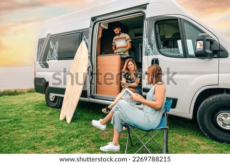 Group of friends traveling in a camper van. Two women talking in front of camper van while young man cooks