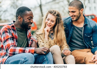 Group Of Friends Two Men And One Woman With Mobile, Sitting On A Bench In Park