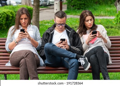 Group of friends two women and one man, sitting on a bench in park separately looking at their phones loosing communication. people using their phones and sending texts as they stand beside each other