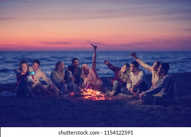 a group of friends using cell phones around the bonfire during autumn beach party drinking beer and having fun - Powered by Shutterstock