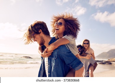 Group of friends walking along the beach, with men giving piggyback ride to girlfriends. Happy young friends enjoying a day at beach. - Shutterstock ID 373830868