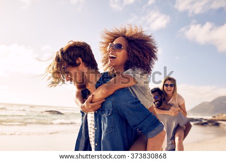 Group of friends walking along the beach, with men giving piggyback ride to girlfriends. Happy young friends enjoying a day at beach.