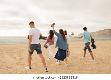 A group of friends walks along the sandy beach, holding cameras and other items, enjoying a fun outing during sunset. The ocean waves gently lap against the shore in the background. - Powered by Shutterstock