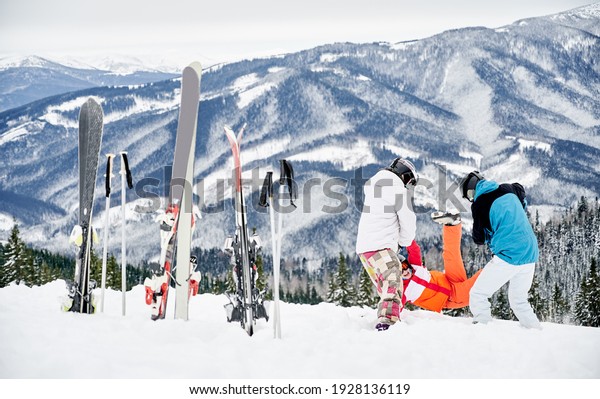 Group of friends\
wearing vivid winter sports suits having fun in snow in mountains.\
Ski poles and skis stuck in snow, people are playing in snow.\
Winter entertainment\
concept