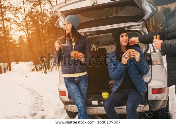 Group friends in\
winter wear on snowy day in the winter forest  having talk and\
drinking coffee in the\
car