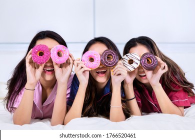 Group Of Funny Teenage Girls Covering Their Eyes With Donuts On The Bed While Having Sleepover Party At Home