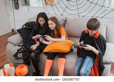 Group Of Girl And Boy Kids Smiling And Dress Up As Halloween Vampire Costume Holding Joystick To Play Halloween Game In Living Room At Home Together