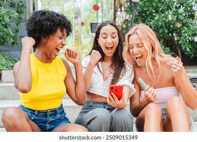 Group of girls looking at smart phone and celebrating winning an online bet. Three succeed young women shouting, cheering, and raising their fists in victory while looking at a cellphone. Achievement - Powered by Shutterstock