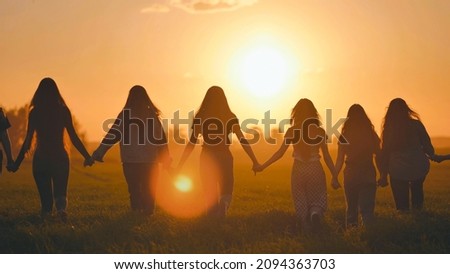 A group of girls walk towards the sun at sunset holding hands.