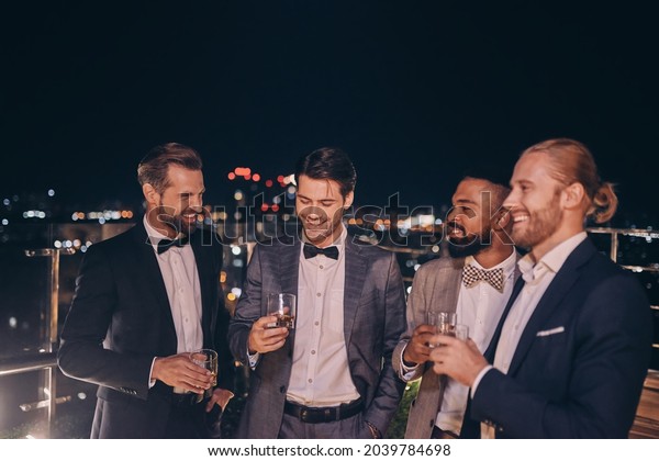 Group of handsome young men in suits and bowties\
drinking whiskey and\
smiling