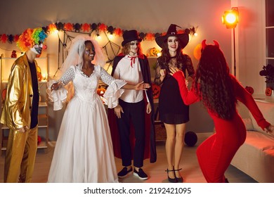 Group Of Happy Adult Friends Meeting At Halloween Fancy Dress Party. Cheerful Young People In Spooky Costumes Of Devil, Dead Bride, Clown, Witch And Vampire Having Fun At Friendly Gathering At Home