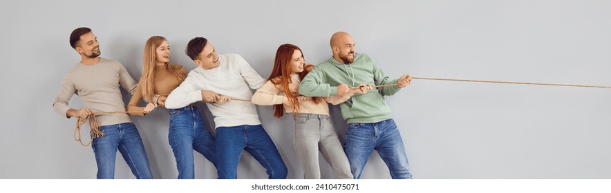 Group of happy adult friends playing tug of war and applying teamwork and unity strategy. Team of cheerful confident people standing together by grey wall pulling rope in one direction. Header, banner - Powered by Shutterstock