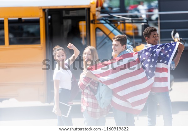 group of happy american teen scholars with usa flag\
in front of school bus