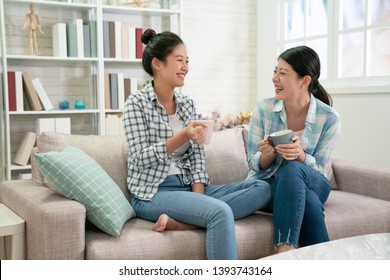 Group Of Happy Asian Female Friends Talking And Laughing Loud Holding Drinks Sitting On Sofa In The Living Room At Home. Young Girls Sisters Gossip Having Fun On Couch With Tea Cups Indoors Apartment