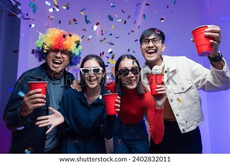 Group of happy Asian friends smiling at party, drinking. Young man and woman having fun at colorful party at home. Wear dark sunglasses. The room was lit up with neon disco lights and paper shoots.