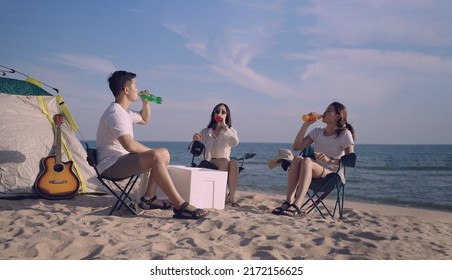 Group Of Happy Asian Young Friends Sitting On Camping Chairs Holding And Drinking Colored Soft Drinks On Beach During Summer Vacation. Young Woman And Man While Cocktail Party Near Tent On Seaside. 