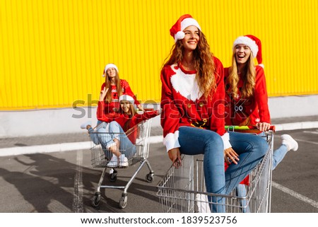 Group of happy cheerful young women in christmas sweaters and santa claus hats walking down city street riding shopping trolleys near shopping mall