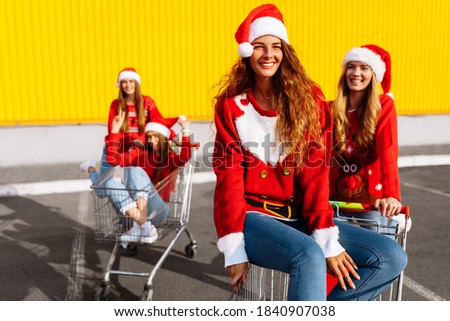 Group of happy cheerful young women in christmas sweaters and santa claus hats walking down city street riding shopping trolleys near shopping mall