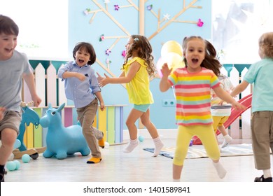 Group Of Happy Children Of Boys And Girls Run In Day Care. Kids Playing In Kindergarten