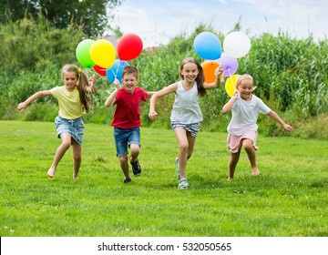 Group Of Happy Children Playfully Running With Multicolored Balloons On Green Meadow

