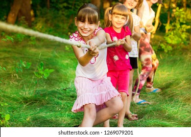 Group Of Happy Children Playing Tug Of War Outside On Grass. Kids Pulling Rope At Park. Summer Camp Fun