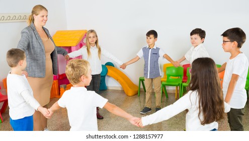 Group Of Happy Children Standing In Circle Holding Hands, Playing With Their Teacher In Classroom During Recess 