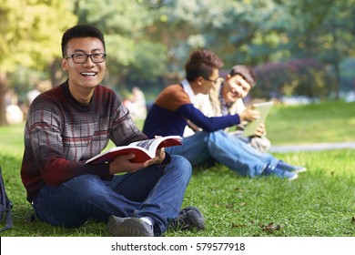 Group Of Happy Chinese College Students  Reading Book On Lawn In Campus