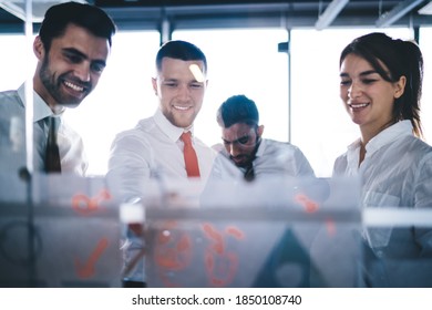 Group Of Happy Coworkers In White Shirts Cheerfully Looking At Business Diagrams Posted On Glass Wall In Modern Conference Room