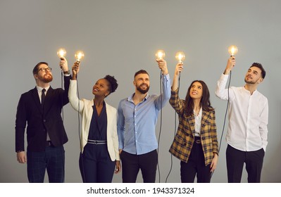 Group of happy creative young diverse business professionals holding glowing light bulbs standing on gray studio background. Innovative thinking, finding solution, people developing own idea concept - Shutterstock ID 1943371324