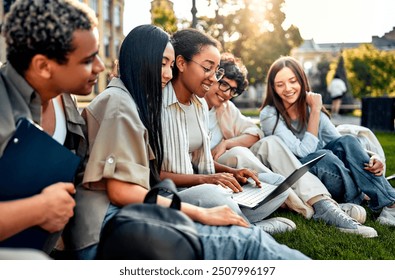 Group of happy diverse students sitting on grass and studying with laptop on university campus. Student life, high school. Motivation and learning.     