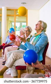 Group Of Happy Elderly Ladies In A Seniors Gym Doing Hand Coordination Exercises Throwing And Catching Brightly Colored Plastic Balls As They Sit In Their Chairs