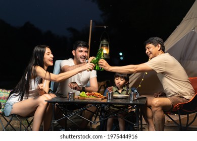 Group Of Happy Family Traveler Enjoyed Camping In The Forest At Night, Caucasian Father, Asian Mother, And Grandfather Sitting At A Folding Table Clinking The Green Bottles Of Beer For Celebration.