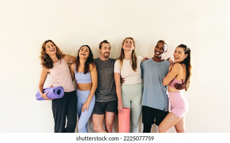 Group of happy fitness friends laughing while standing against a wall with yoga mats. Multicultural people attending a yoga class together. Sporty people working out in a community fitness studio. - Powered by Shutterstock