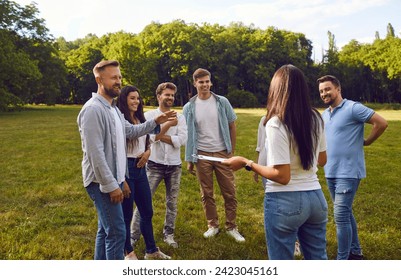 Group of happy friends or corporate coworkers standing on grass in summer park on weekend and listening to manager with clipboard listing their tasks and explaining rules of outdoor team building game - Powered by Shutterstock