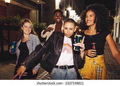 Group of happy friends dancing in the city at night. Four vibrant young people holding beer cans while walking together in the street. Friends having a good time on a weekend night. - Shutterstock ID 2107198811