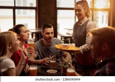 Group of happy friends drinking beer in a tavern while waitress is serving them nacho chips.